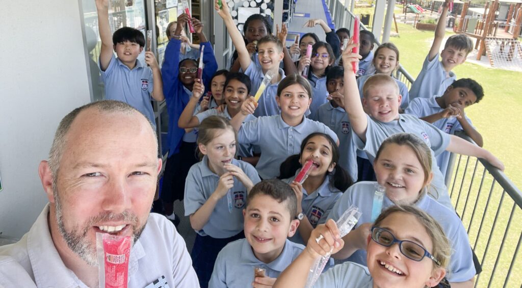 Students in uniform sharing a fun moment with their teacher, enjoying popsicles and smiling at the camera. Hearing the Bible in our Heart Language.